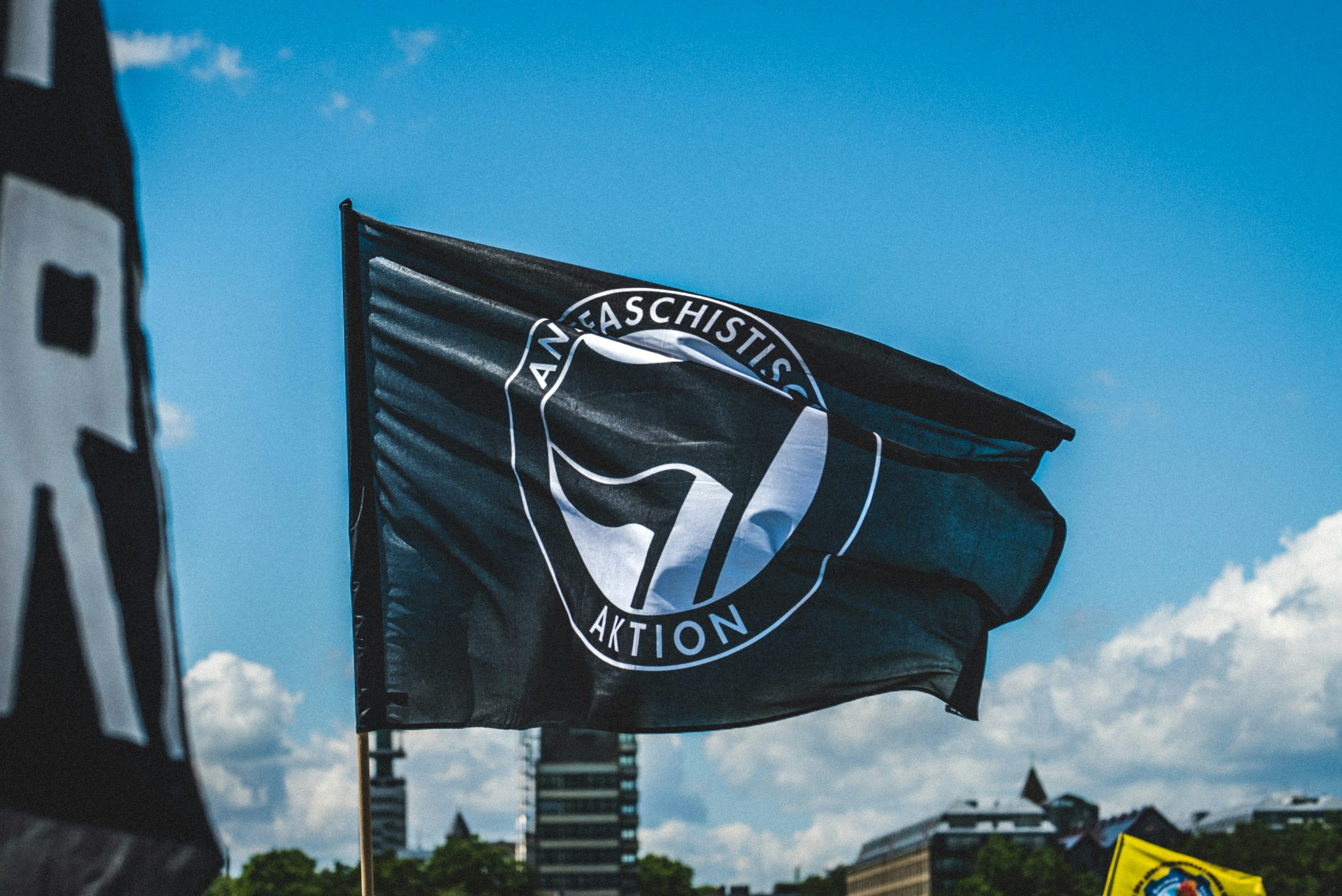 Schwarz weiße Flagge mit Logo "Antifaschistische Aktion" auf blauem Himmel, anscheinend Teil einer Demonstration (man sieht n ur die Flagge und ein paar Häuser im Hintergrund)